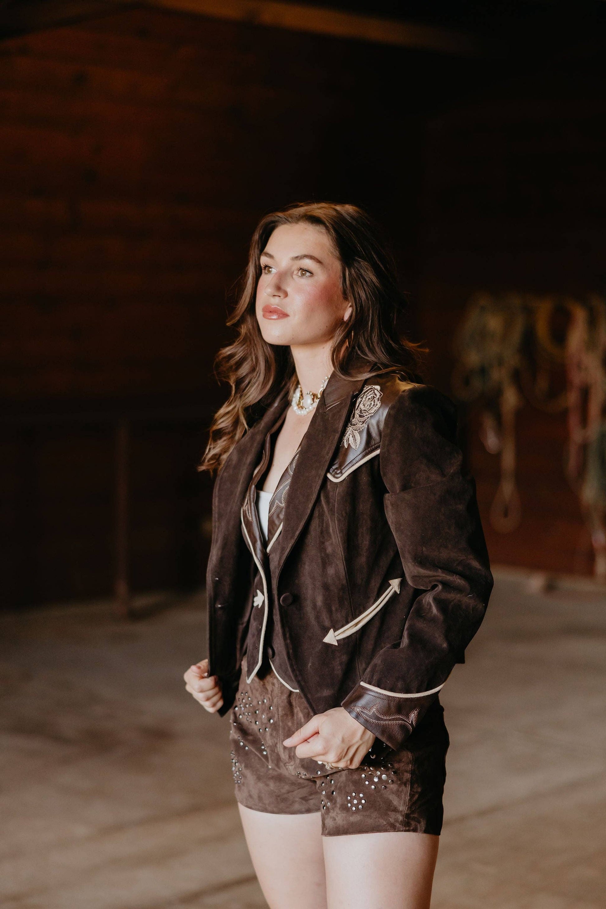 Woman wearing a dark brown blazer with decorative elements in a dimly lit room.