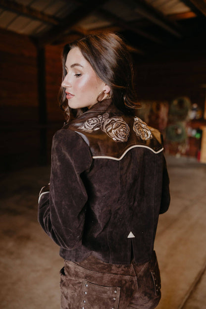 Woman wearing a brown suede jacket with floral embroidery in a dimly lit room.