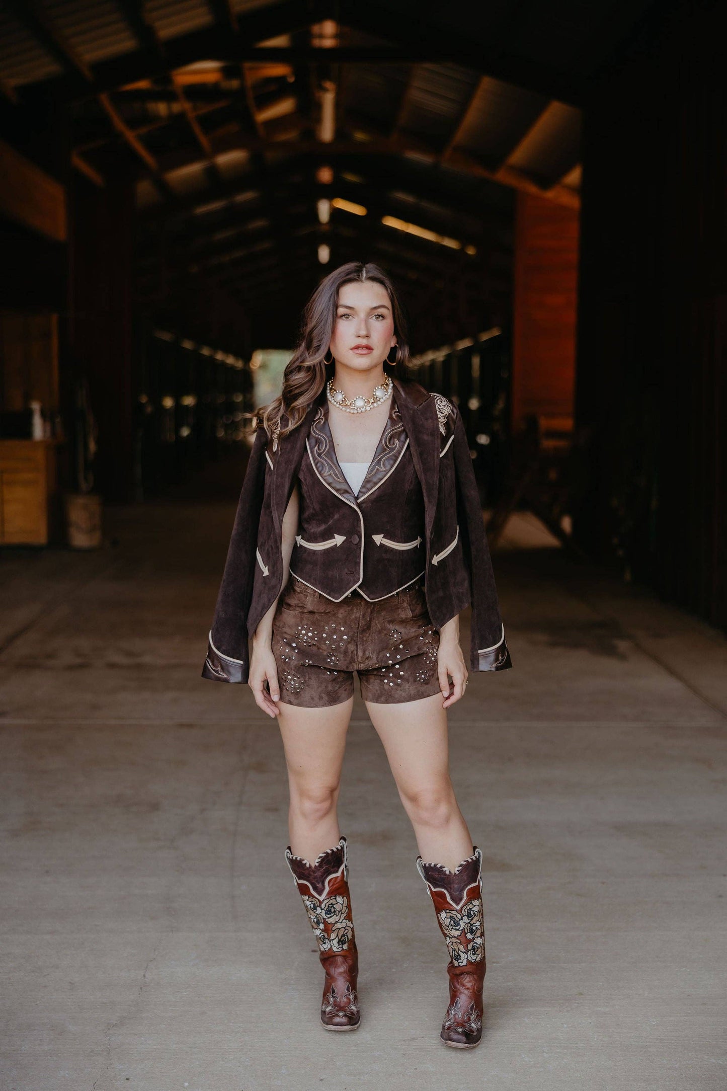 Woman in a brown western-style outfit with boots standing in a dimly lit barn.